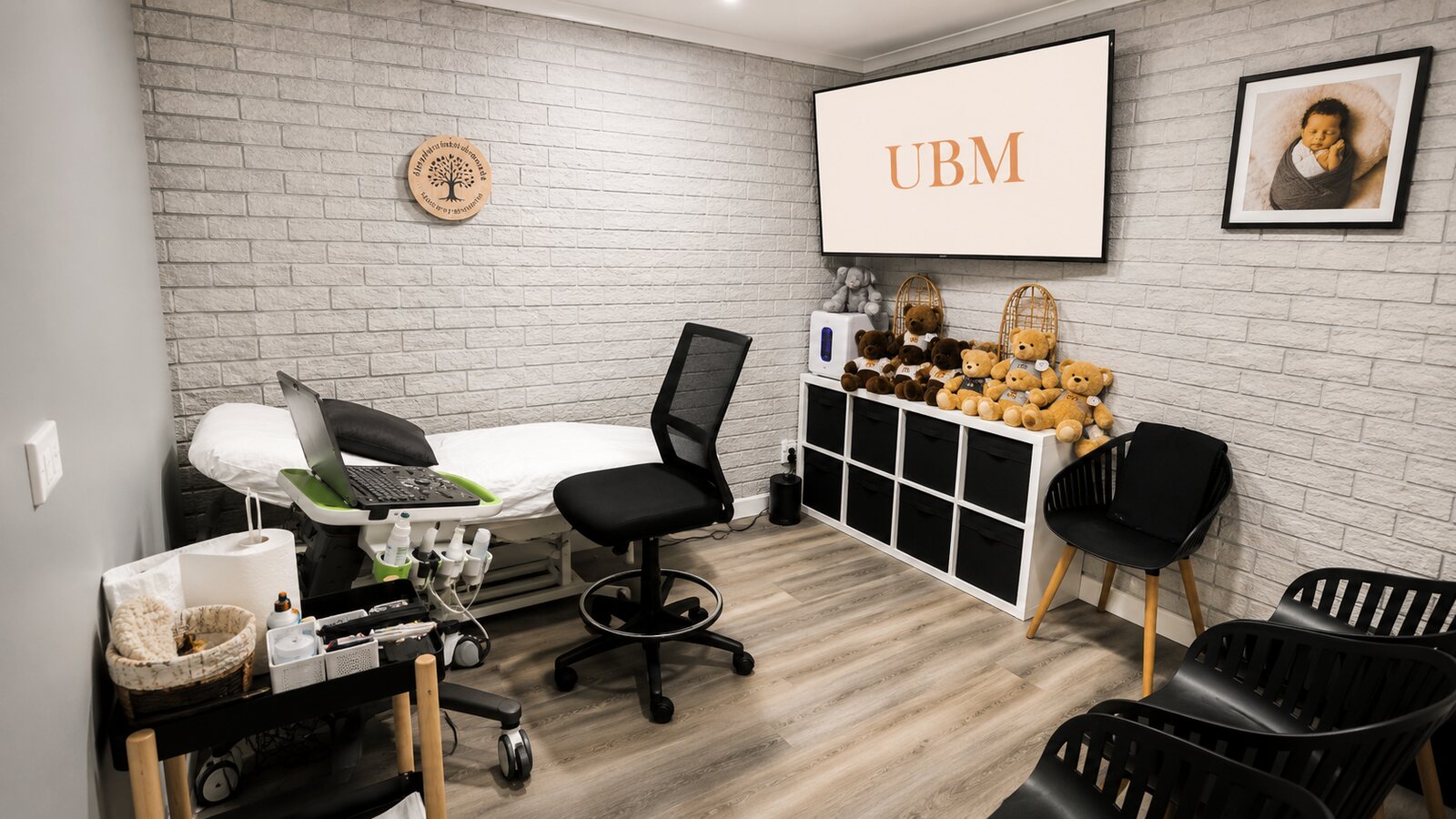 A scanning room at the UBM Scans Menai clinic: an examination bed and ultrasound machine beside a large wall-mounted screen, with a shelf of teddy bears and seating for family against the white-brick wall
