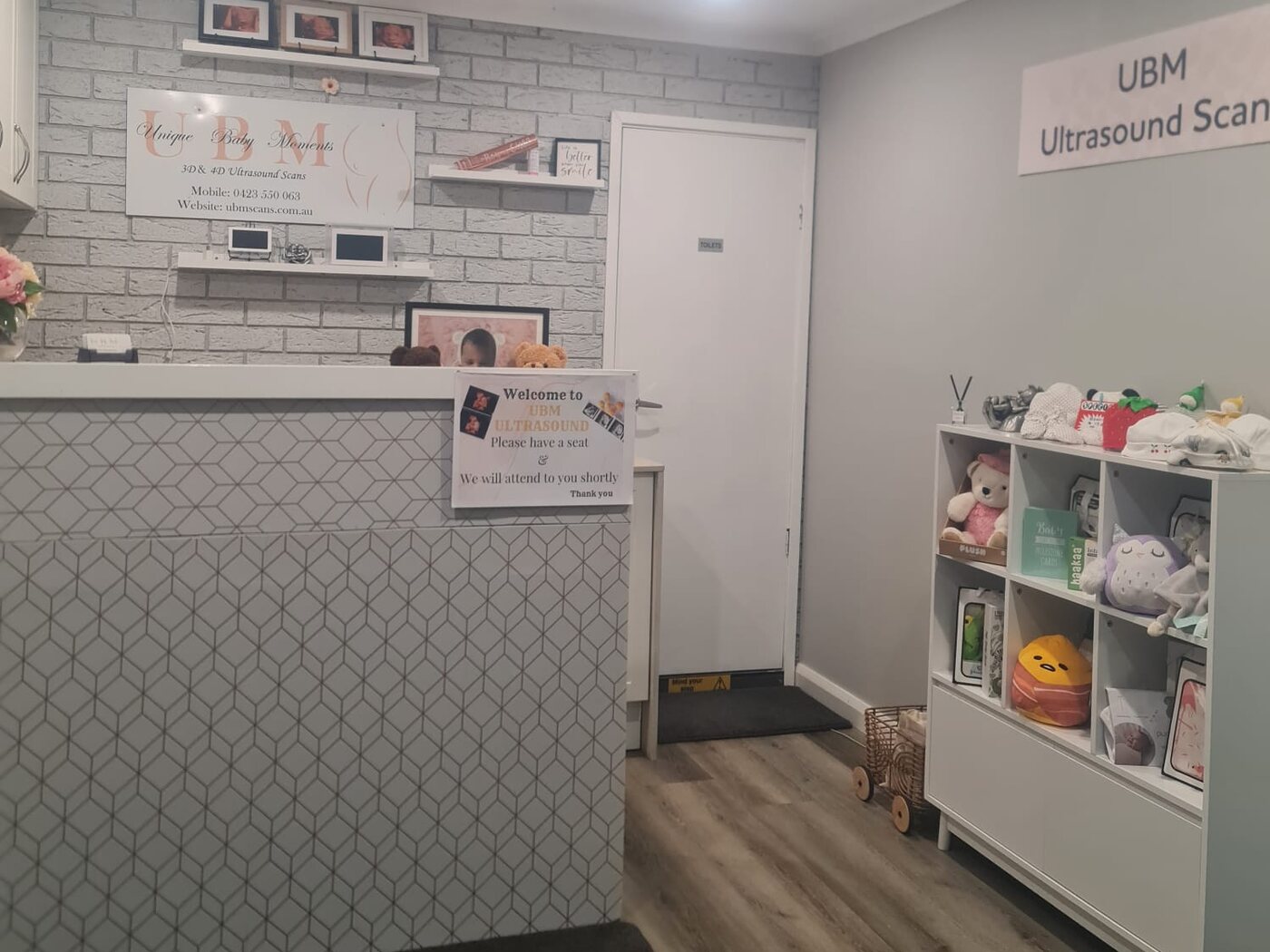 The UBM Scans reception in Menai: a tiled front desk beneath the Unique Baby Moments sign, with scan prints framed on the brick wall and a shelf of keepsakes beside the waiting area