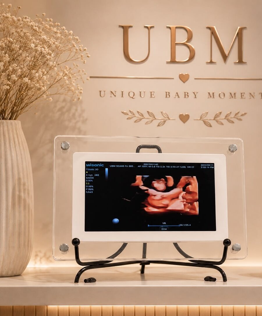 A 4D ultrasound image displayed in a clear acrylic UBM Scans keepsake frame on a black stand, on the clinic reception bench beneath the gold Unique Baby Moments wall sign